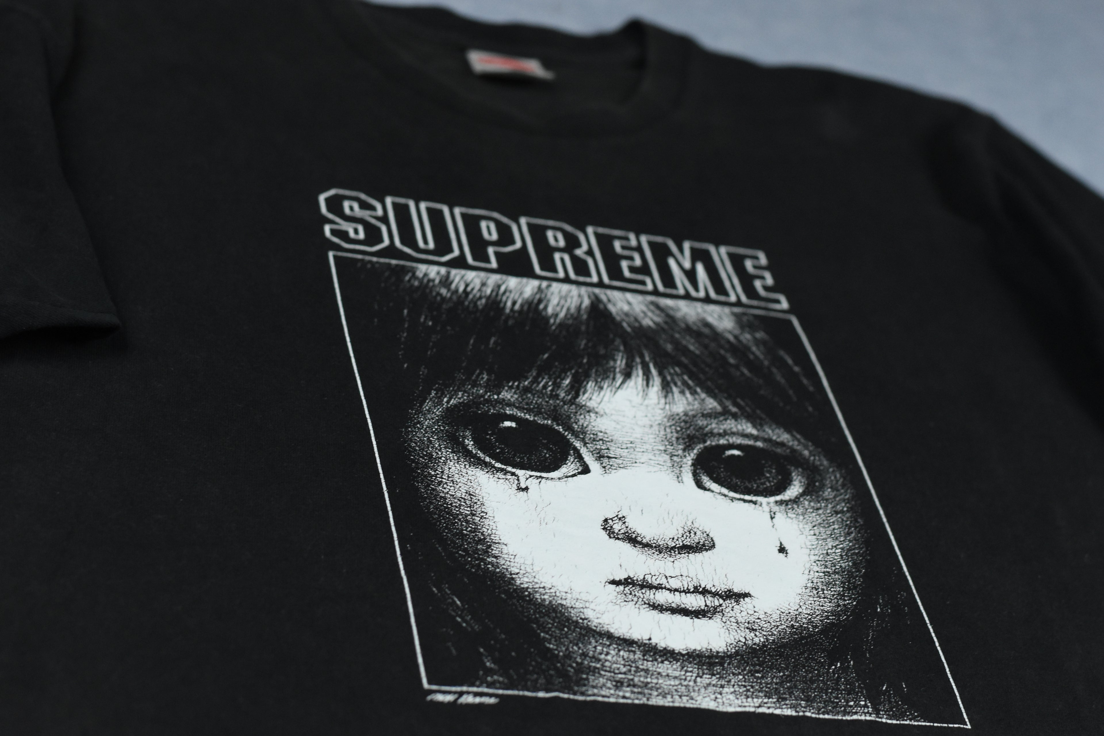 Black t-shirt with a graphic of a doll's face and 'Supreme' logo.